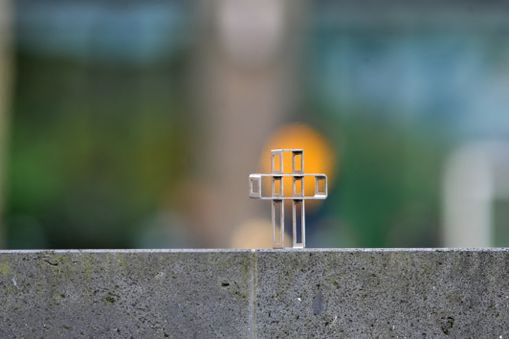 Metallkreuz durch das man schauen kann, gelber Punkt direkt dahinter. Das Kreuz steht auf eienr Mauer.